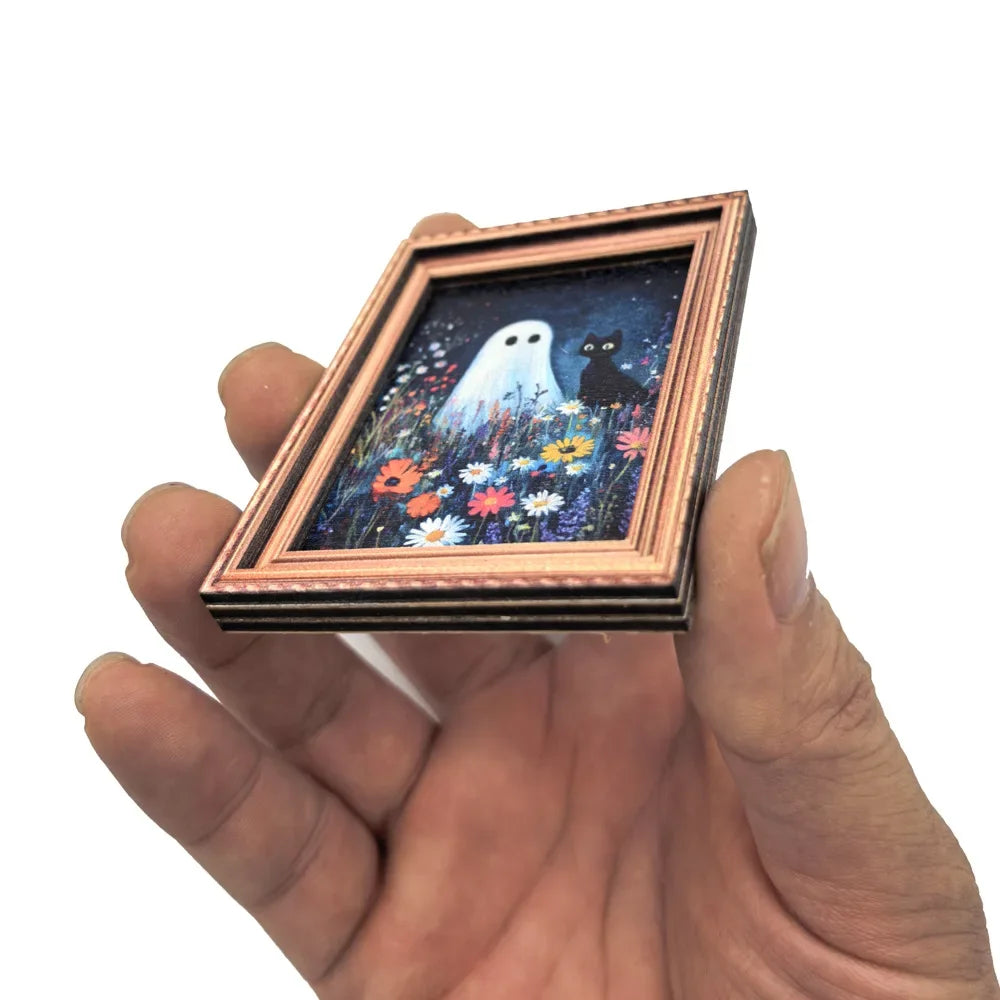 A hand holds a framed picture featuring a white ghost and a black cat among colorful flowers.