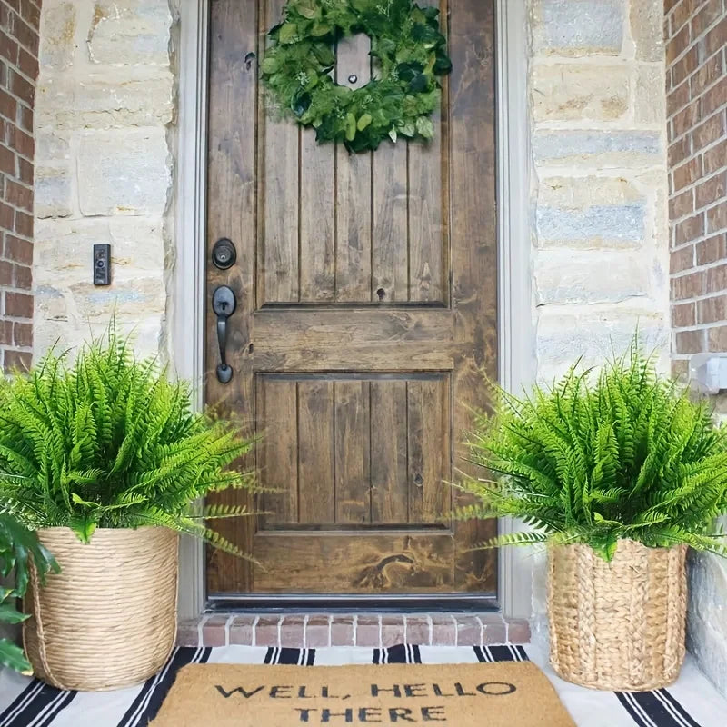 Two potted artificial Boston fern bushes flank a rustic wooden door decorated with a wreath.