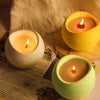 A yellow silicone arc candle jar with a lit candle sits on a wooden surface next to some paper.