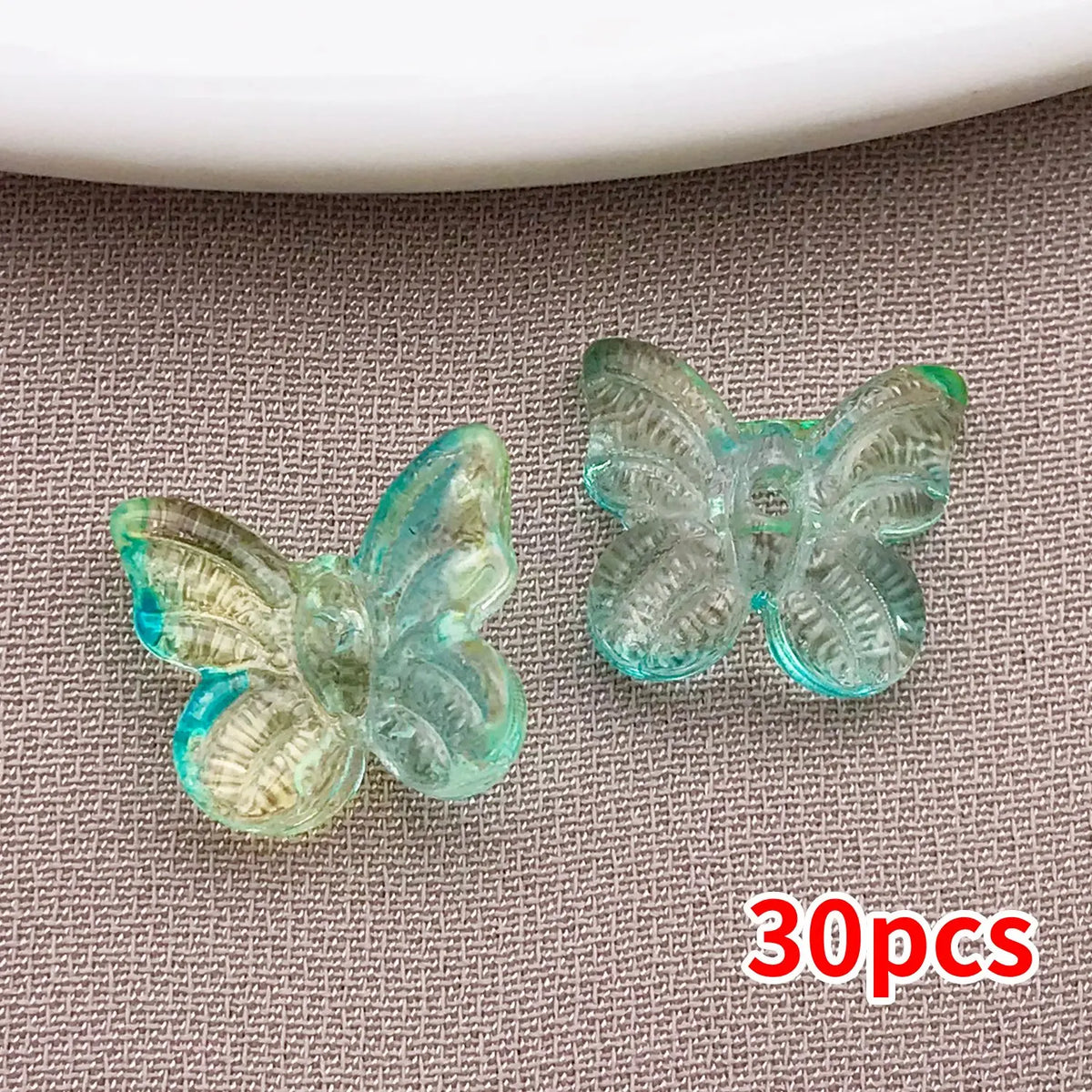 Un juego de 30 cuentas de mariposa de vidrio transparente y azul claro, exhibidas sobre una superficie gris texturizada.