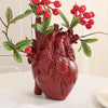 A realistic, deep red heart sculpture made of resin with green leaves and red berries on a white surface.