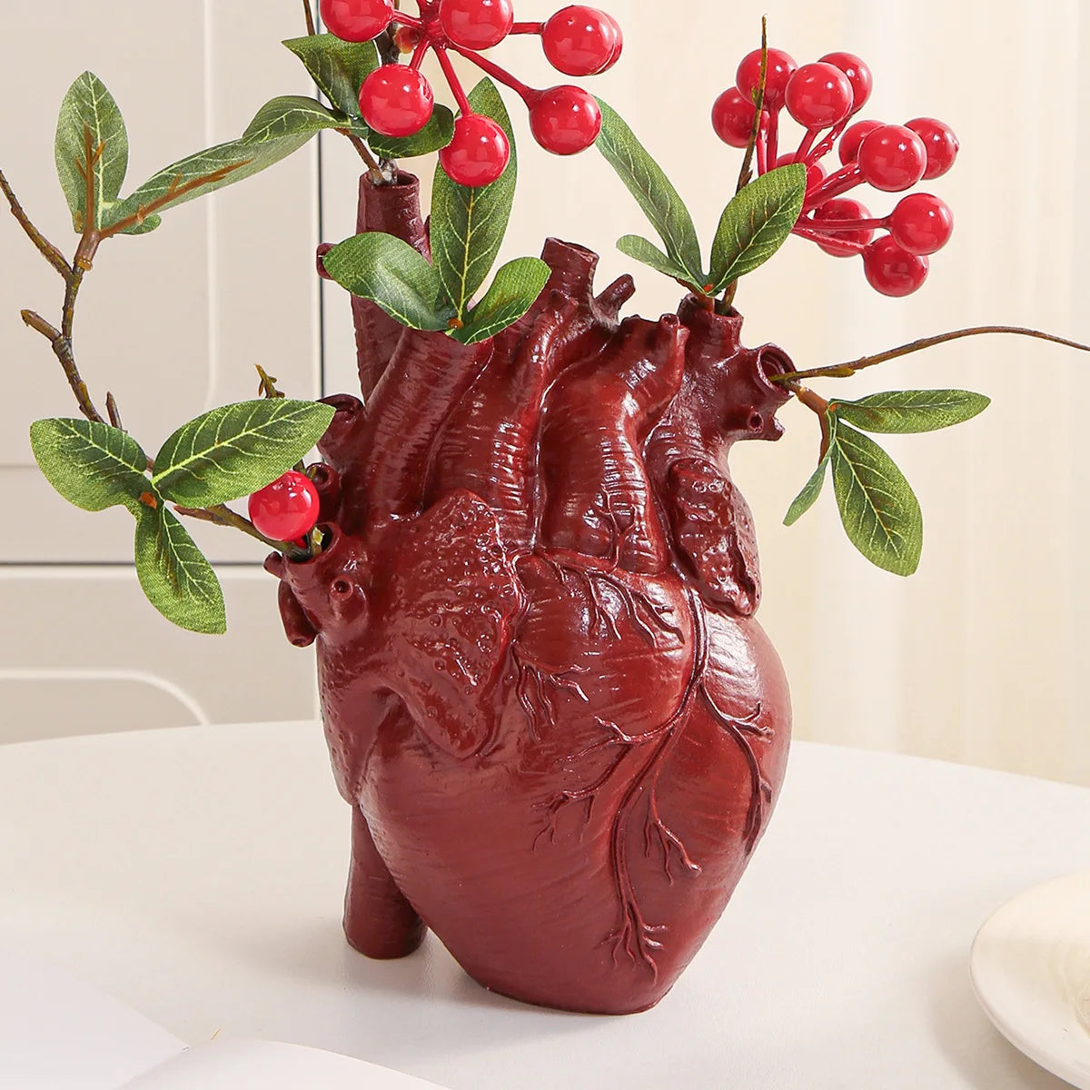 A realistic, deep red heart sculpture made of resin with green leaves and red berries on a white surface.