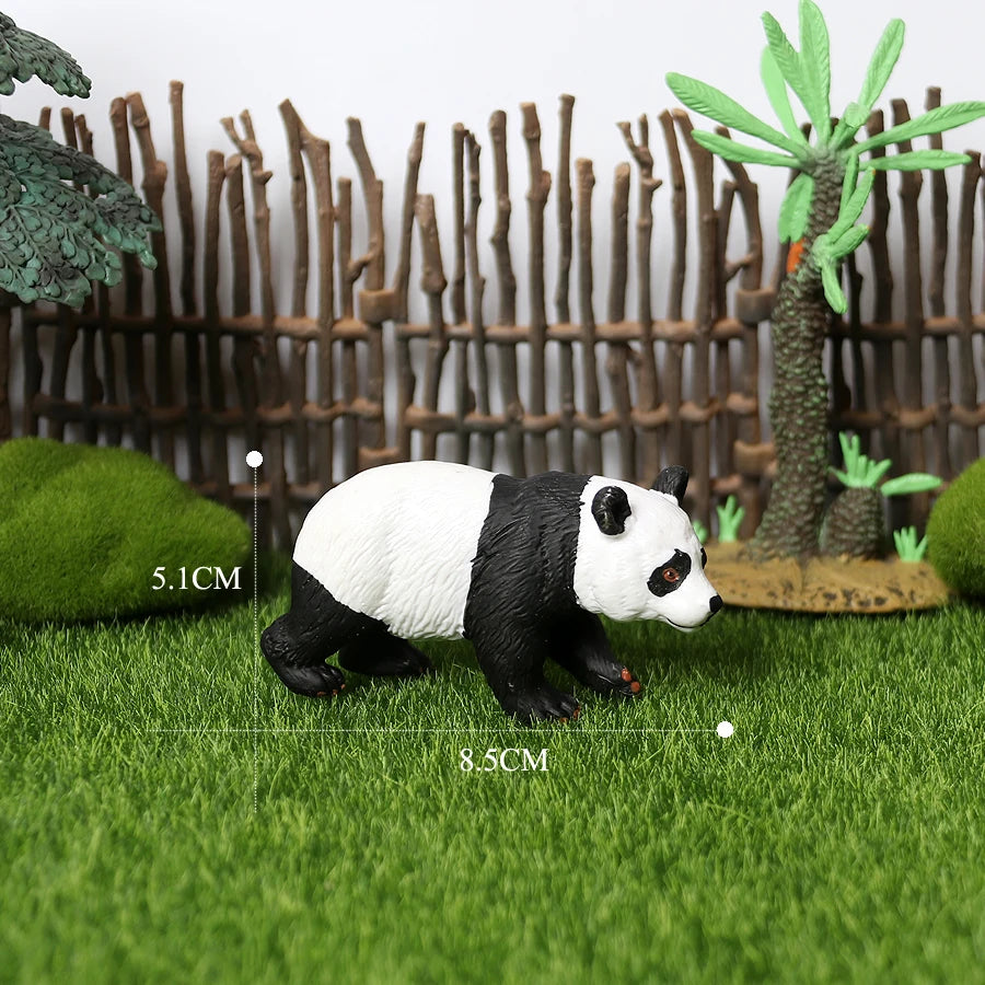 A realistic black and white panda figurine standing on green grass with a bamboo fence and palm plants in the background.