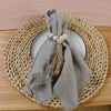 A stack of ten handcrafted fringed cotton napkins in gray are displayed on a round woven mat.