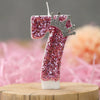 A pink sequin number seven candle with a silver crown topper displayed on a wooden surface.