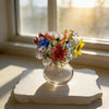 Handcrafted blue glass flower sculpture with colorful petals displayed on a clear glass base near a window.