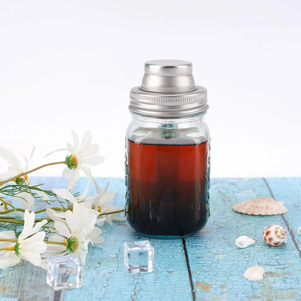 A glass jar filled with dark amber liquid sits on a wooden surface next to white flowers and ice cubes.