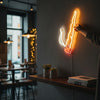Une main tient une décoration de cigarette néon illuminée sur un mur blanc.