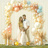 A floral arch made of light pink, cream, and gold balloons decorated with dried pampas grass and a couple.
