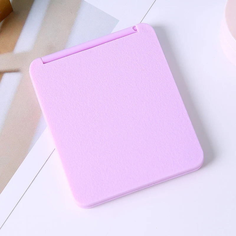 A pink compact foldable travel makeup mirror with a smooth finish is shown on a white surface.