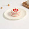 A stack of pink and white layered cake on a white paper plate with red berries, next to a dried floral element.