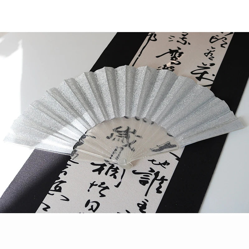 A silver glitter folding fan with transparent ribs is displayed against a dark background featuring Japanese calligraphy.