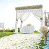 A wedding or party setup featuring a white draped archway decorated with flowers and surrounded by chairs and a field of whit
