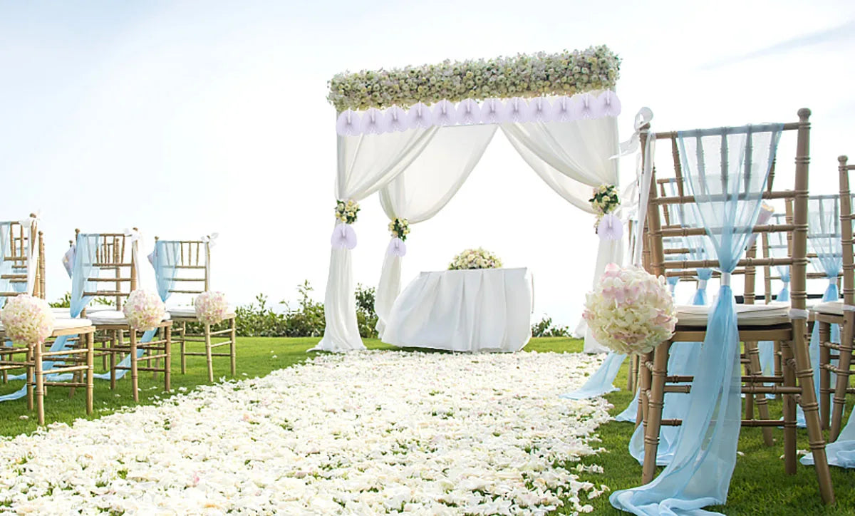 A wedding or party setup featuring a white draped archway decorated with flowers and surrounded by chairs and a field of whit
