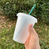 A hand holds a clear plastic cold cup with a green straw outdoors in a grassy area.