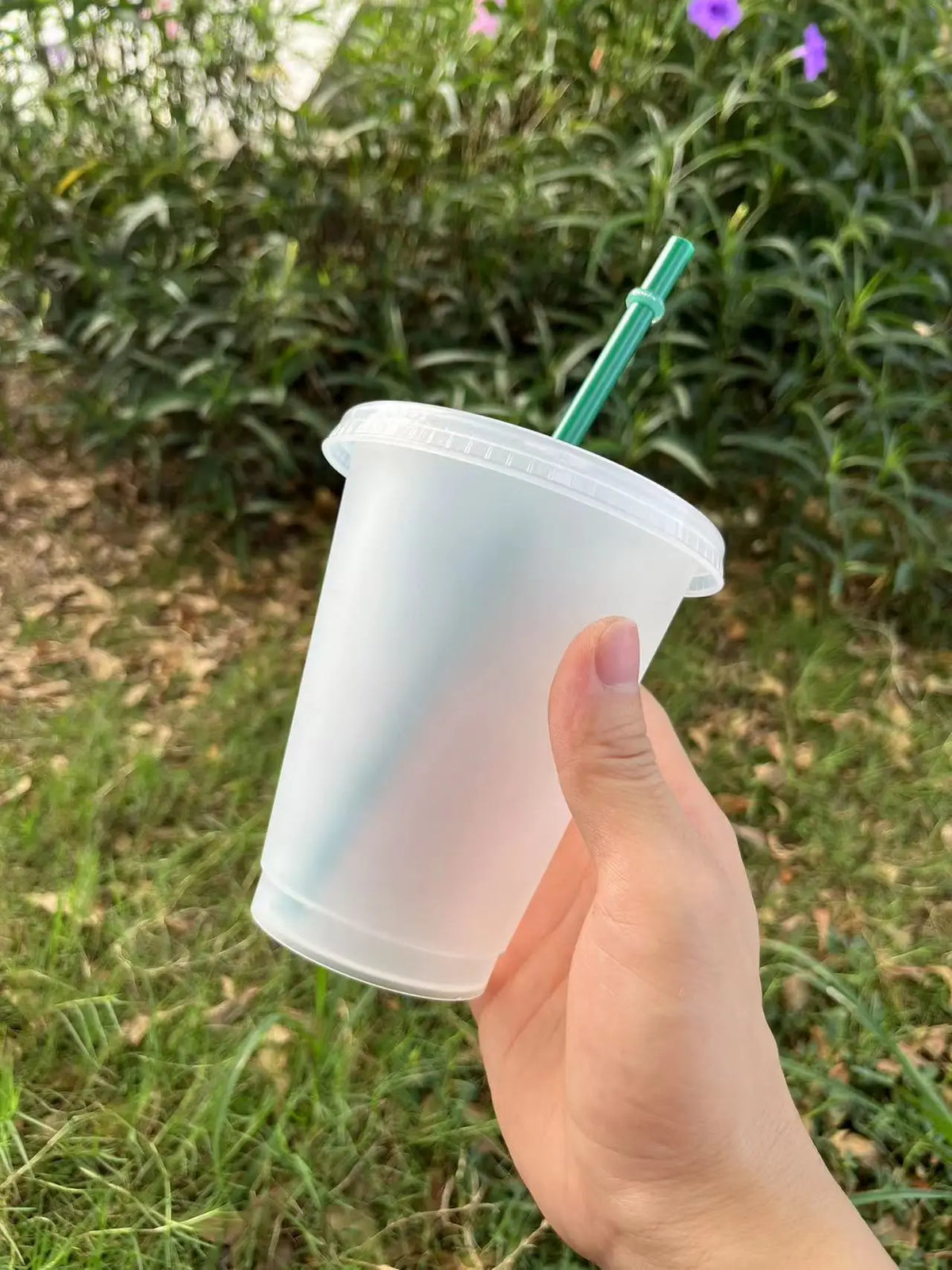 A hand holds a clear plastic cold cup with a green straw outdoors in a grassy area.