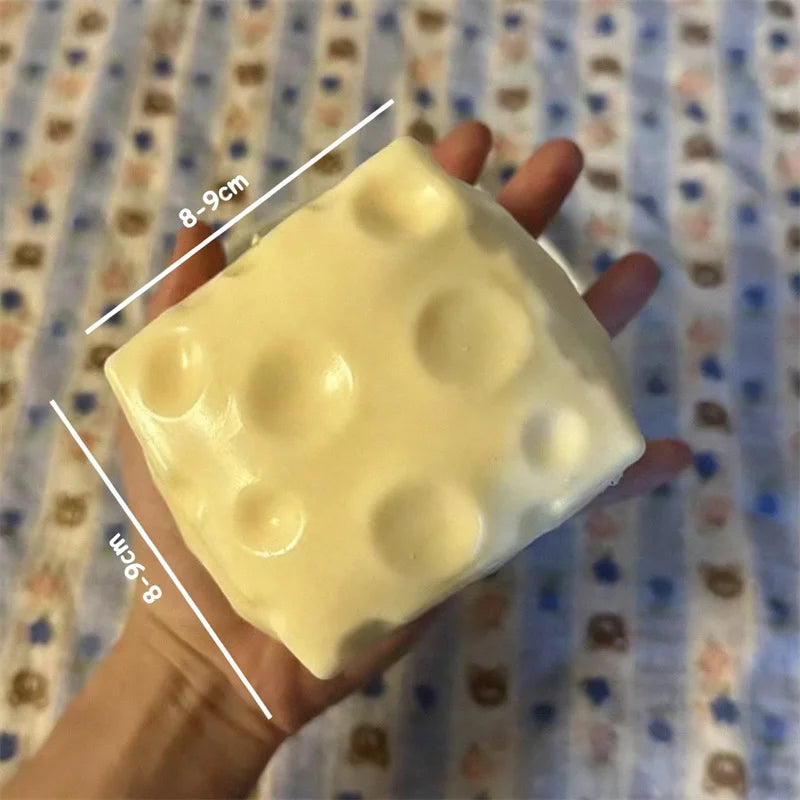 A hand holds a large, pale yellow cheese cube stress ball with visible dimples.