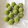A collection of thirty velvet green Christmas baubles in various sizes and textures displayed on a white fabric.