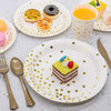 A white plate with gold polka dots holds a slice of cake decorated with colorful frosting and fruit, next to a yellow drink i