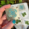 A hand holds a small, square metal flower press with a turquoise and gold pressed flower inside.