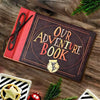 A vintage kraft scrapbook album titled Our Adventure Book resting on a wooden surface with greenery and a star-shaped item.