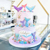 A tiered cake decorated in purple and blue mermaid tail shapes, seashells, and sea stars.