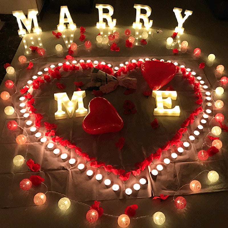 A heart-shaped display made of warm white LED alphabet letters spelling 'MARRY ME' surrounded by red and white lights.