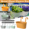 A set of metal plant flower pots in orange, green, and gray hanging planters displayed against a metal fence.