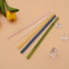 A collection of five reusable borosilicate glass straws in various pastel and dark colors laid diagonally on a beige surface.