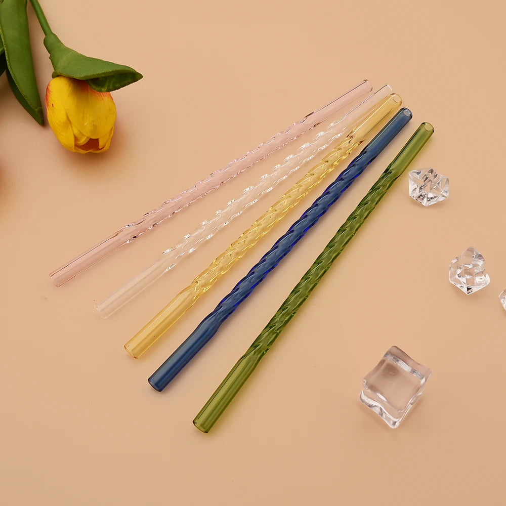 A collection of five reusable borosilicate glass straws in various pastel and dark colors laid diagonally on a beige surface.