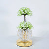 A small potted tree of white flowers in a clear glass vase on a gold base.