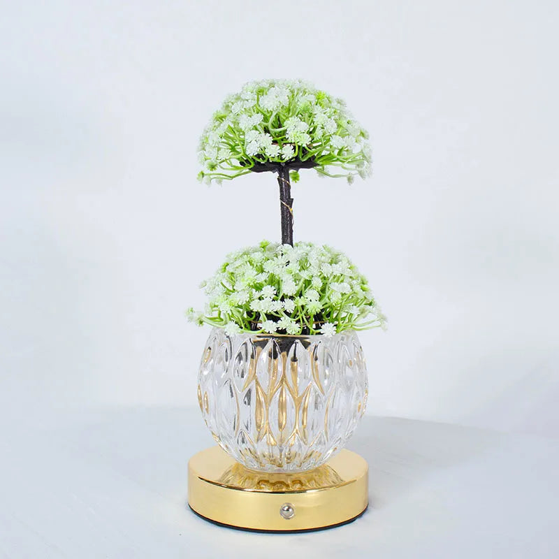 A small potted tree of white flowers in a clear glass vase on a gold base.