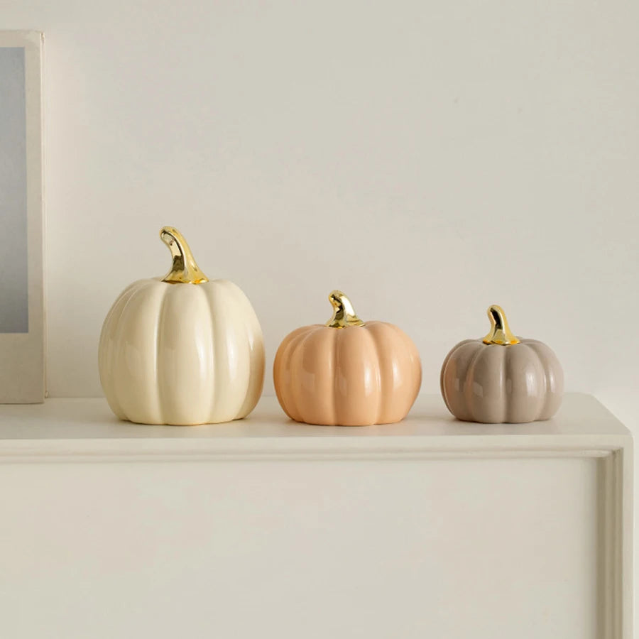 Three ceramic pumpkin figurines in cream, peach, and gray with gold accents displayed on a white shelf.
