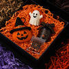 An orange pumpkin, a white ghost figure, and a black witch hat are arranged on crinkle paper filler for Halloween treats.