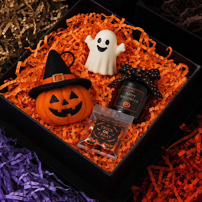 An orange pumpkin, a white ghost, and a jar of Halloween candy are arranged on crinkle paper shred filler for decor.