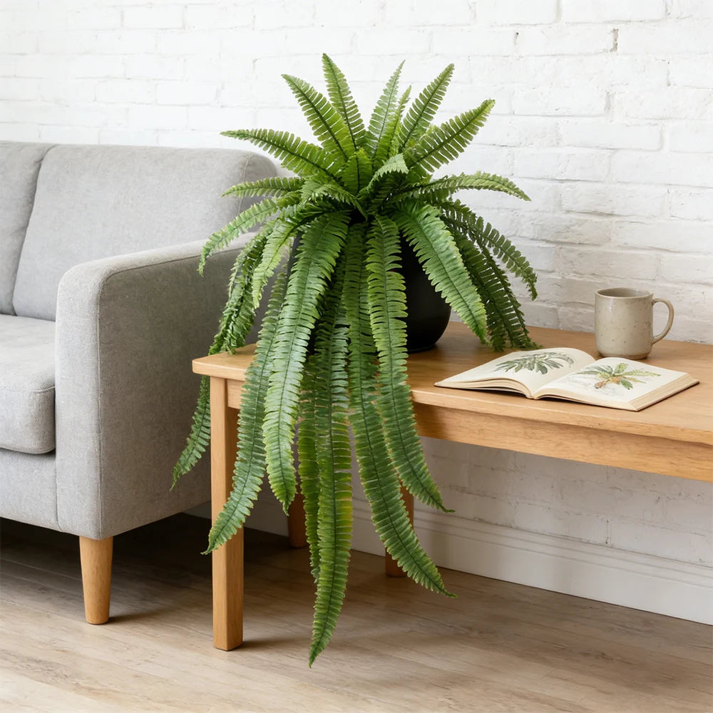 A large artificial Persian fern hanging vine in a black pot rests on a wooden table next to an open book.