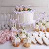 A white cake decorated with pink floral elements and purple ribbon bows, surrounded by small cupcake toppers.