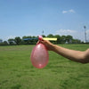 A hand is holding a pink balloon with a yellow ribbon against a bright blue sky and green field.