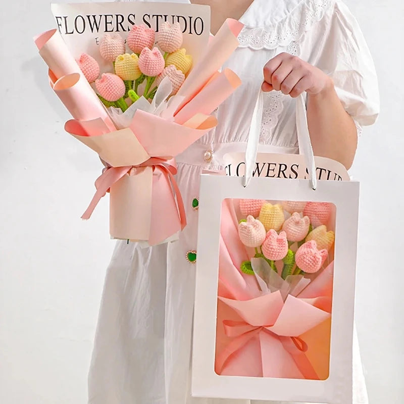 A person holds a bouquet of handmade crochet tulip flowers wrapped in pink paper and a white gift bag.