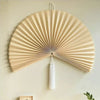 Handwoven bamboo wall fan decor with a light beige color and white tassel hanging on a wall.