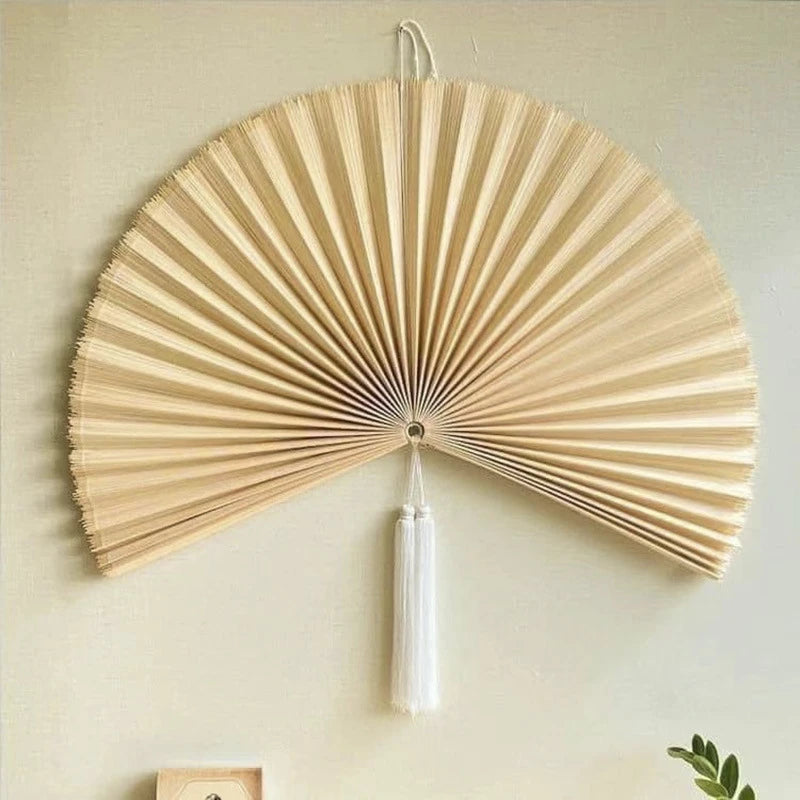 Handwoven bamboo wall fan decor with a light beige color and white tassel hanging on a wall.