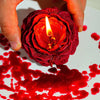 Eine Hand hält eine tiefrote Rosenkerze mit brennender Flamme, die Schatten auf eine mit rolicher Flüssigkeit befleckte Oberf