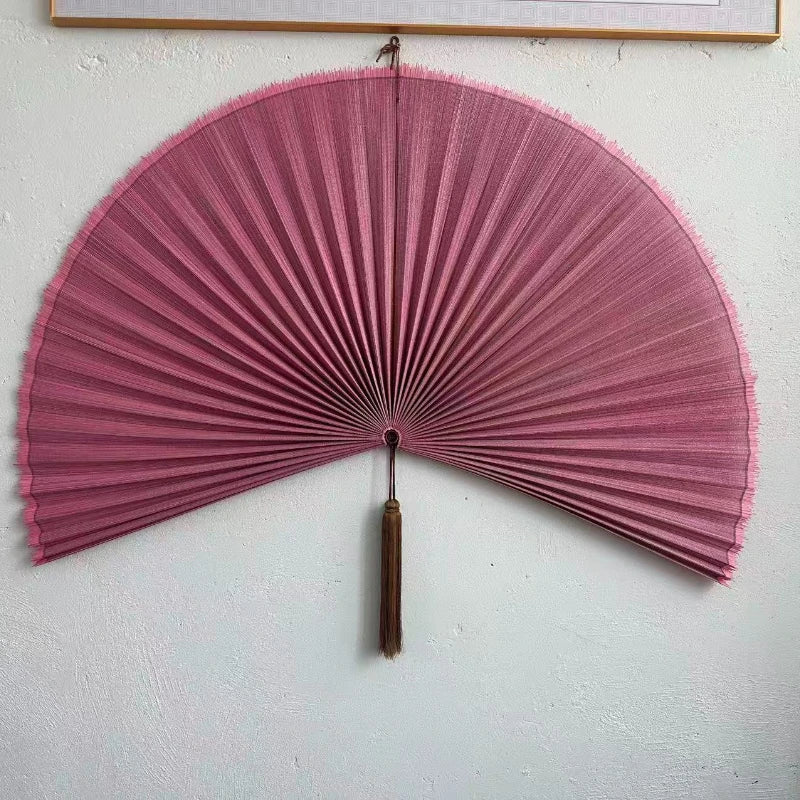 A pink handwoven bamboo wall fan decor with a tassel mounted on a white wall.