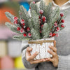 A hand holds a bouquet of frosted pine needles and red berries wrapped in twine.