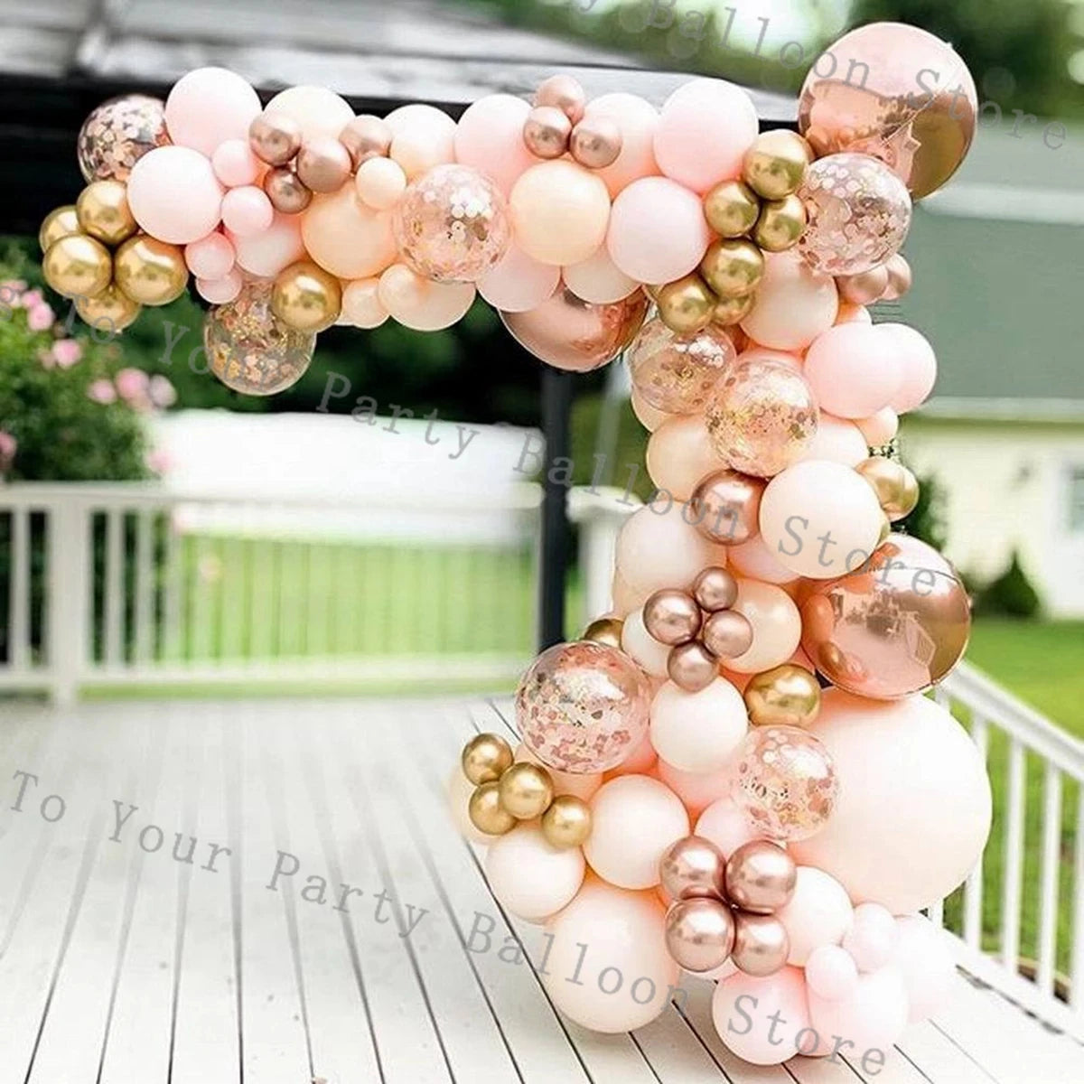 A balloon garland arch made of pink and gold confetti balloons displayed outdoors on a wooden deck.