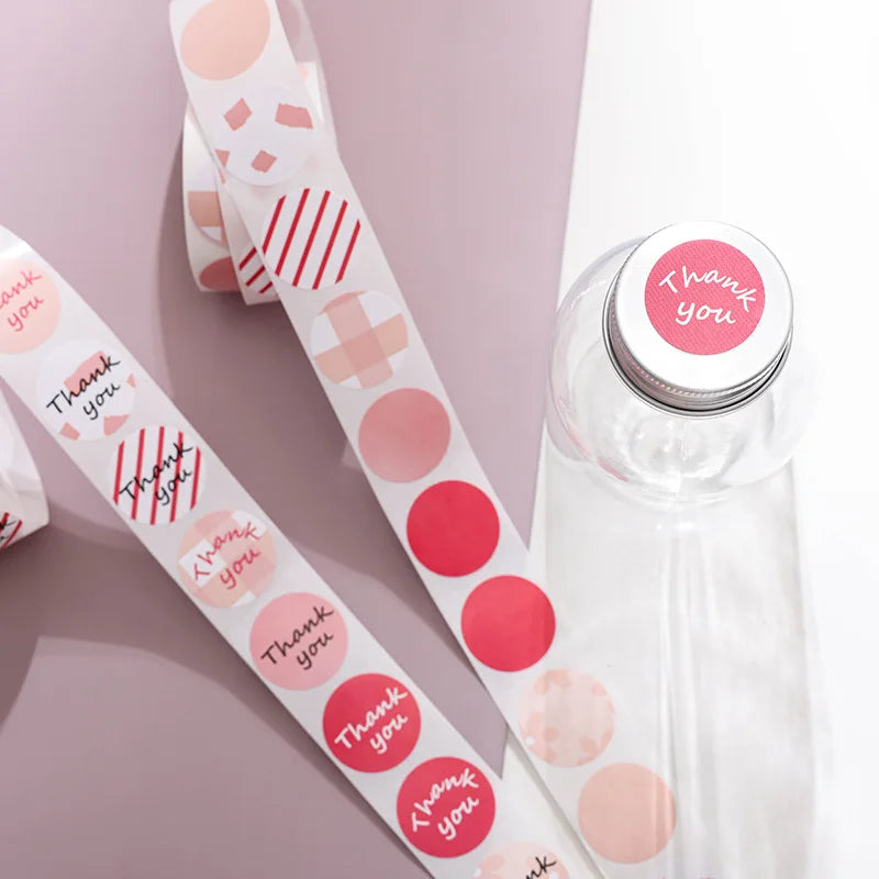 Eine Rolle runder pinker und roter Dankeschön-Sticker neben einer klaren Flasche.