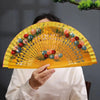 A hand-painted decorative folding fan with yellow petals and floral designs is held by a person.