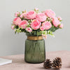 A bouquet of pink silk peony roses in a green glass vase with small green foliage.