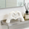 A white heart shape sculpture made of hands rests on a metal shelf in front of a tiled backsplash.