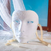 A white ancient style masquerade mask with intricate floral designs is displayed on a white cloth.
